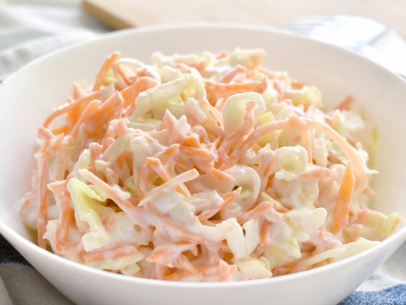 Draufsicht, schräg: Ein Coleslaw in einer weißen Schüssel auf einem blau-weiß kariiertem Küchentuch.