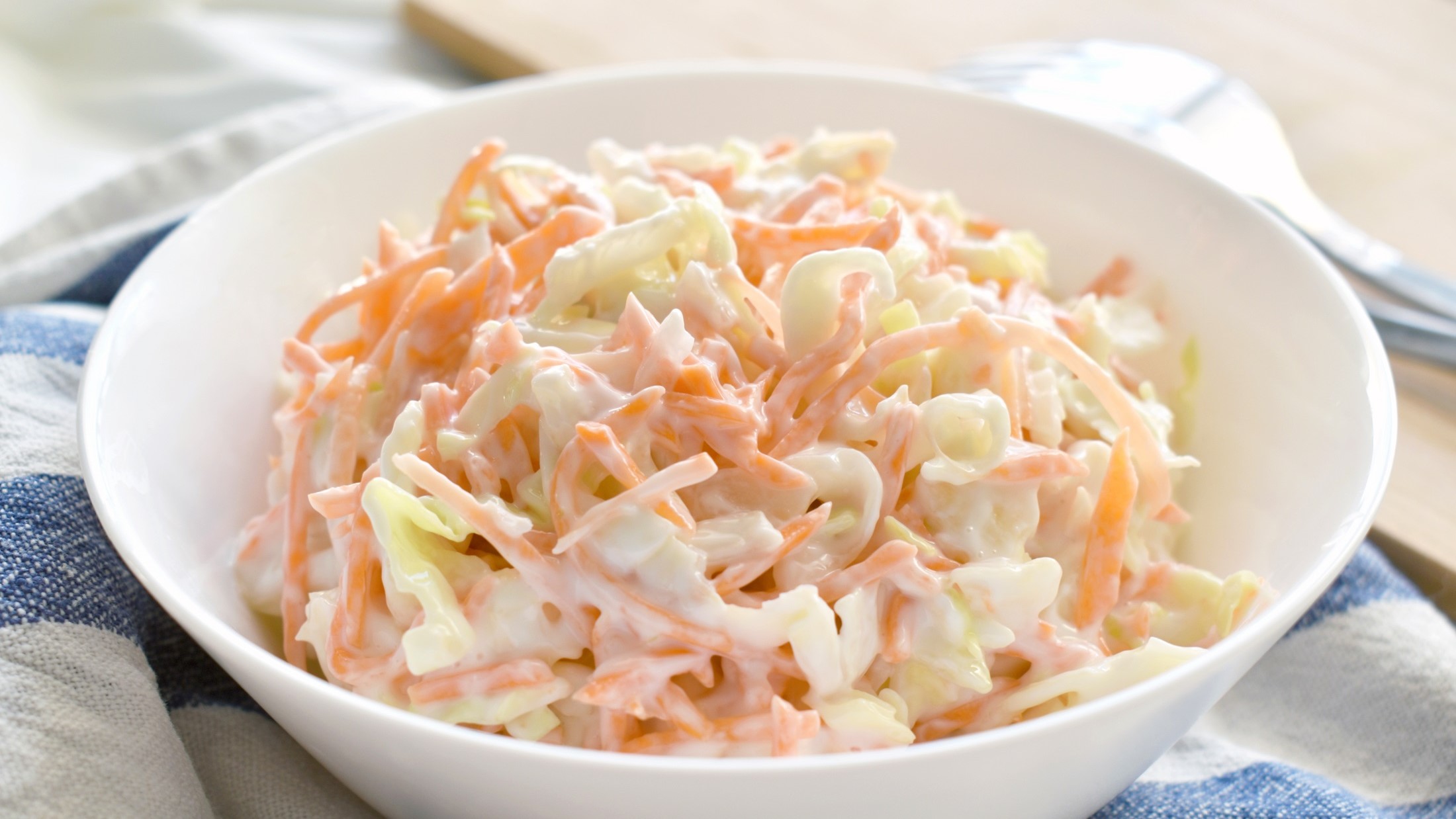 Draufsicht, schräg: Ein Coleslaw in einer weißen Schüssel auf einem blau-weiß kariiertem Küchentuch.