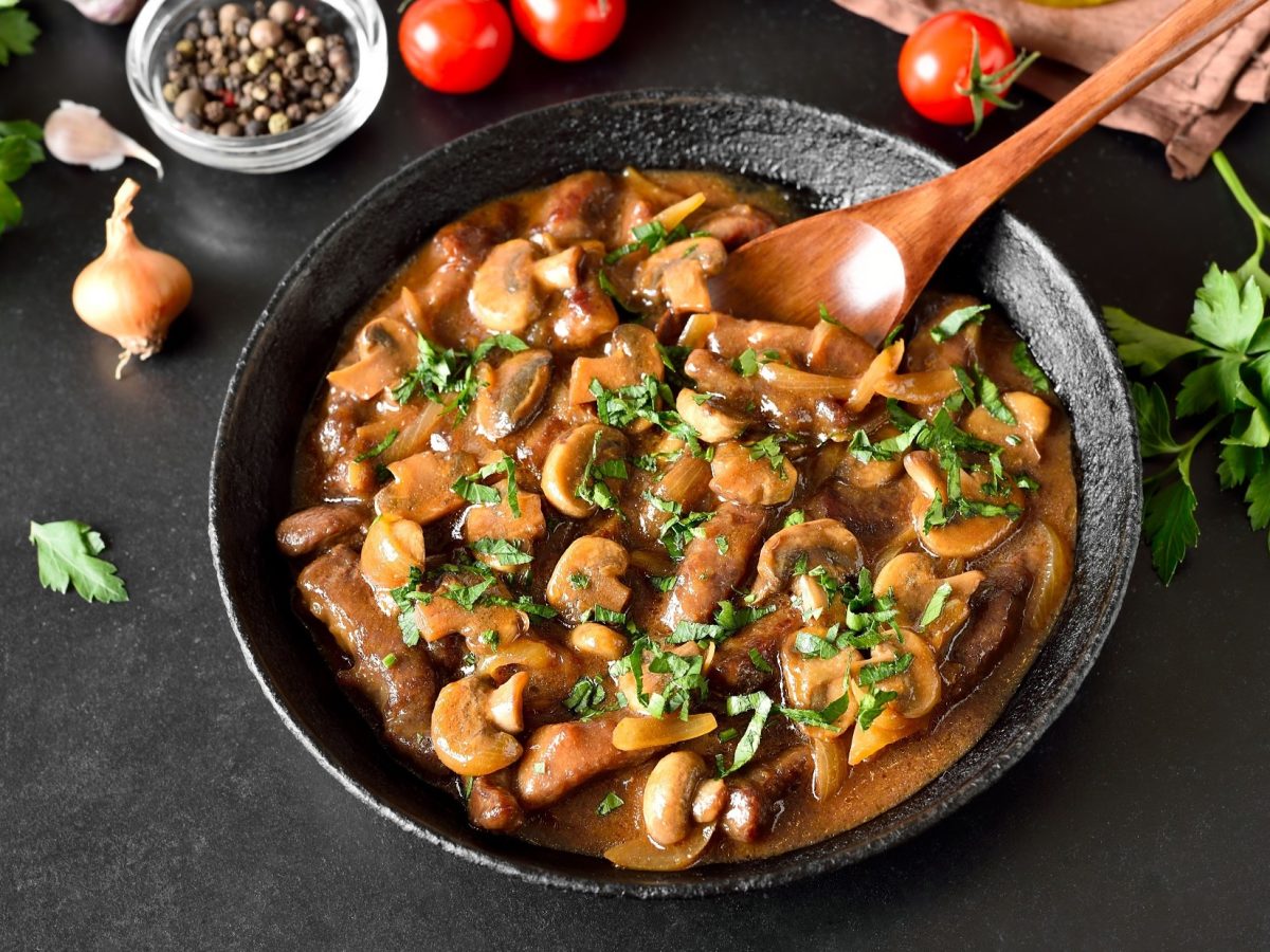 Draufsicht: Eine Pfanne Gulasch mit Pilzen auf schwarzem Untergrund. Drum herum liegen frische Petersilie, Tomaten, Zwiebeln und ein Schälchen mit Salz.