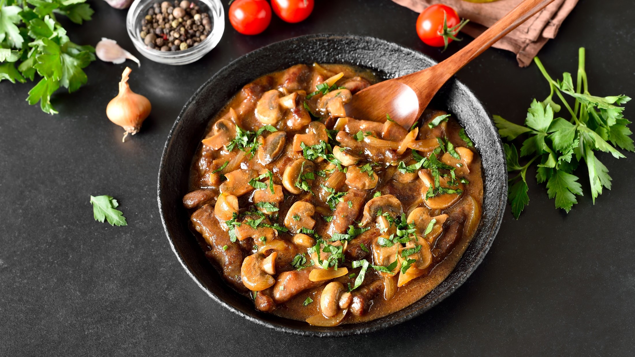 Draufsicht: Eine Pfanne Gulasch mit Pilzen auf schwarzem Untergrund. Drum herum liegen frische Petersilie, Tomaten, Zwiebeln und ein Schälchen mit Salz.