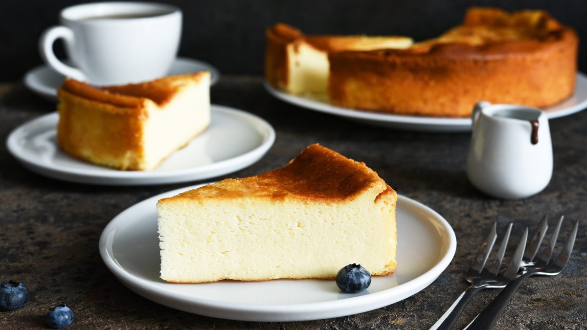 Frontal: Mehrere Stücke Käsekuchen auf drei Tellern auf einem rustikalen Untergrund aus Holz. Um sie herum stehen eine weiße Tasse auf einem Teller und ein Milchkännchen / einfrieren