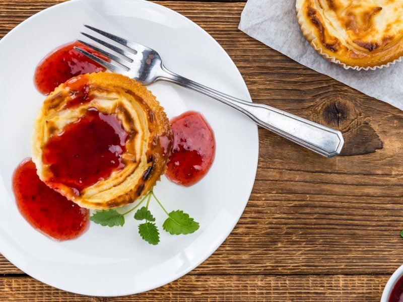 Draufsicht: Ein weißer Teller mit einem Käsekuchen-Muffin mit Fruchtsauce steht auf einem Holztisch. Der Muffin ist mit frischen Kräutern garniert. Drum herum ist ein weiterer Muffin.