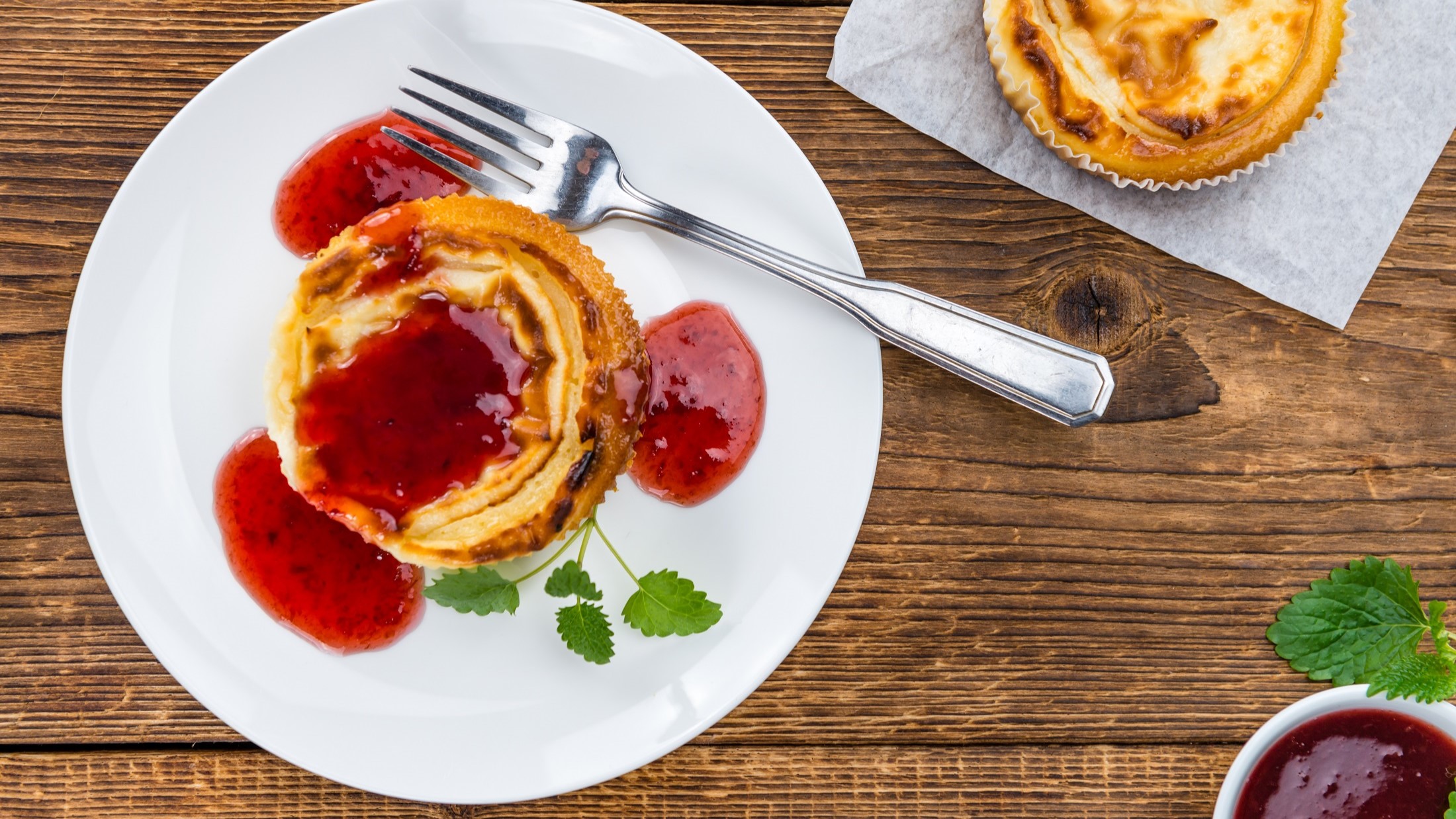 Draufsicht: Ein weißer Teller mit einem Käsekuchen-Muffin mit Fruchtsauce steht auf einem Holztisch. Der Muffin ist mit frischen Kräutern garniert. Drum herum ist ein weiterer Muffin.