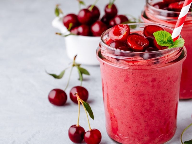 Frontal: Zwei Kirsch-Bananen-Smoothie ohne Milch in einem Glas, in dem je ein weiß-roter Strohhalm steckt. Drum herum und aus dem Smoothie liegen frische, halbierte Kirschen.