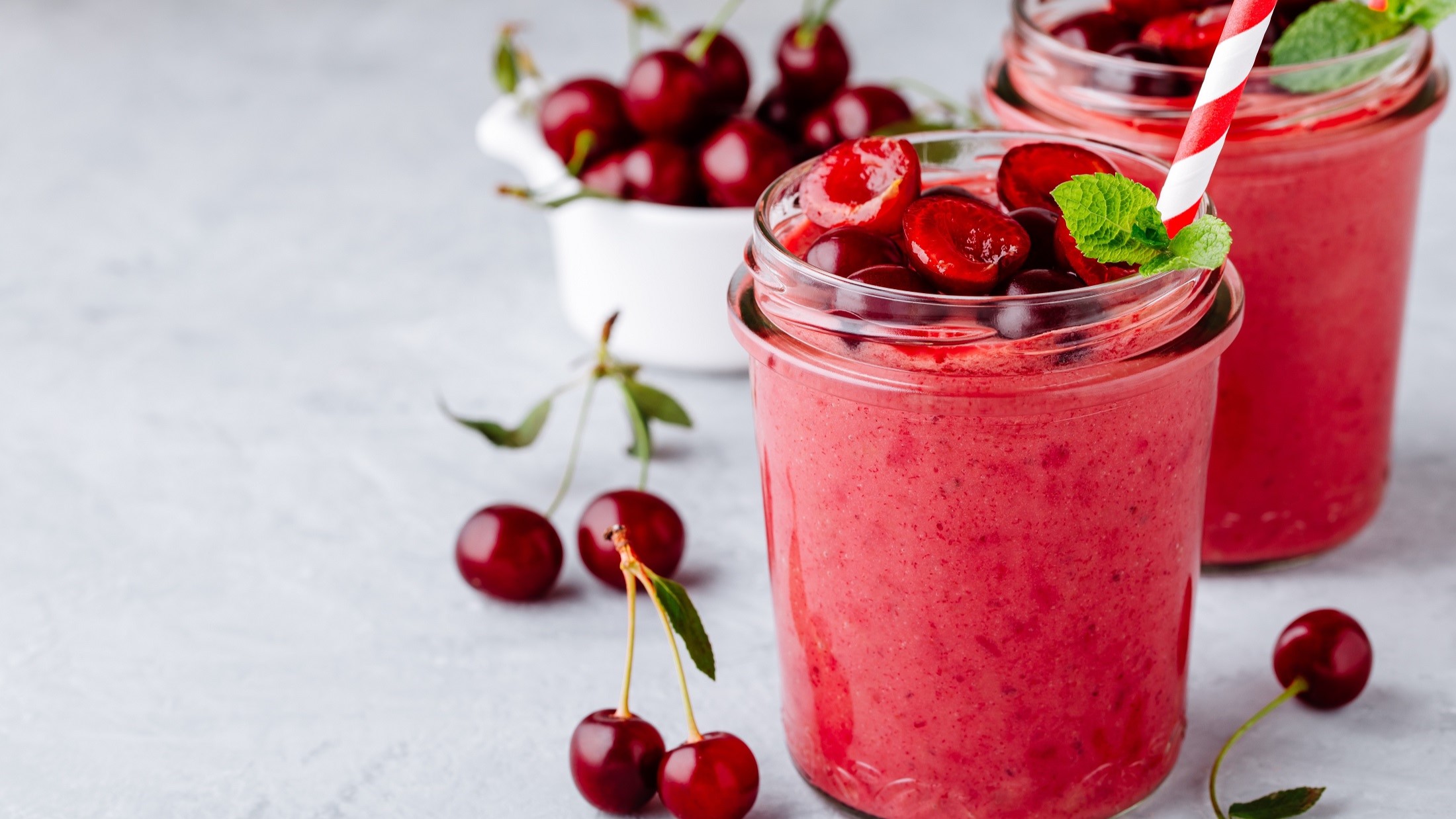 Frontal: Zwei Kirsch-Bananen-Smoothie ohne Milch in einem Glas, in dem je ein weiß-roter Strohhalm steckt. Drum herum und aus dem Smoothie liegen frische, halbierte Kirschen.