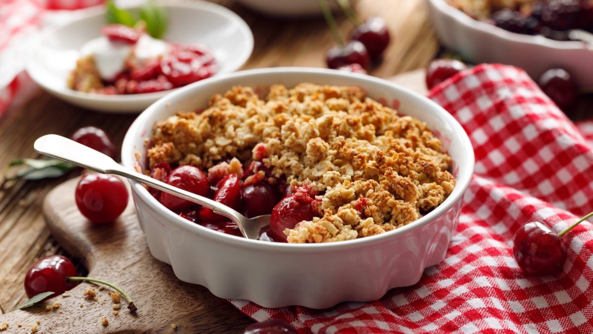 Draufsicht, schräg; Ein runder Kirsch-Crumble mit Streusel in einer runden, weißen Form. Die Form liegt auf einem rot-weiß-kariertem Tuch, dass auf einem Holztisch liegt. Drum herum sind eine kleine weiße Form sowie frische Kirschen.