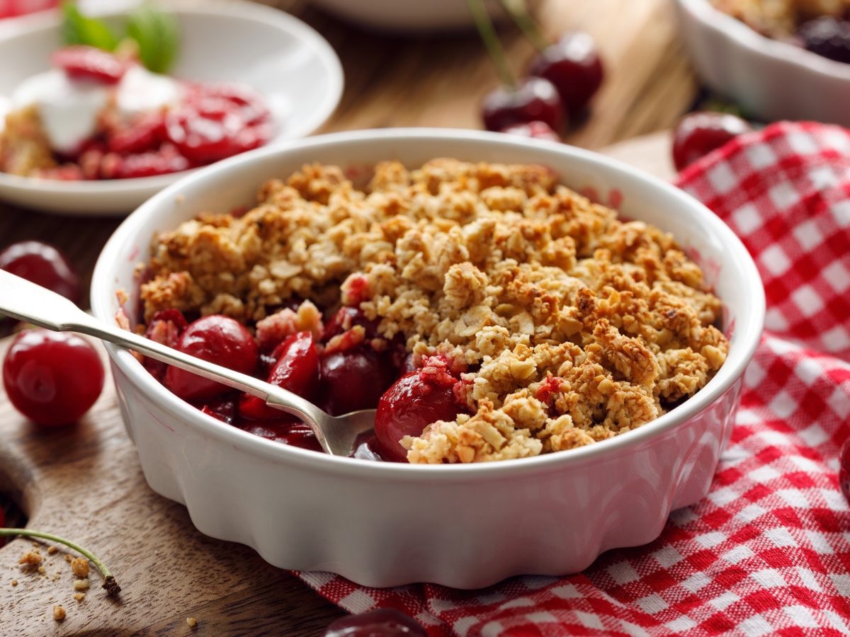 Draufsicht, schräg; Ein runder Kirsch-Crumble mit Streusel in einer runden, weißen Form. Die Form liegt auf einem rot-weiß-kariertem Tuch, dass auf einem Holztisch liegt. Drum herum sind eine kleine weiße Form sowie frische Kirschen.