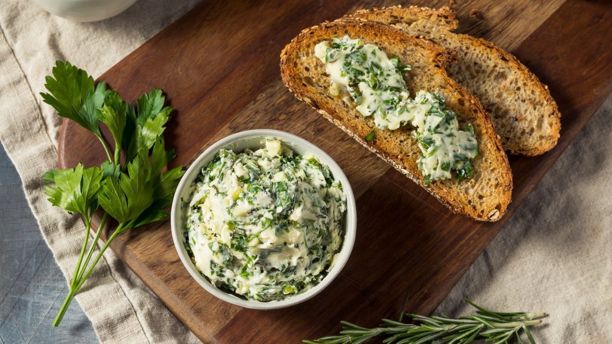 Draufblich: Eine weiße runde Schüssel Kräuterbutter steht auf einem Untergrund aus Holz. drum herum liegen frische Kräuter und zwei Brote mit Kräuterbutter / einfrieren.