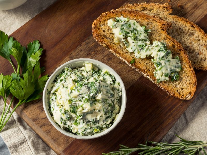 Draufblich: Eine weiße runde Schüssel Kräuterbutter steht auf einem Untergrund aus Holz. drum herum liegen frische Kräuter und zwei Brote mit Kräuterbutter / einfrieren.