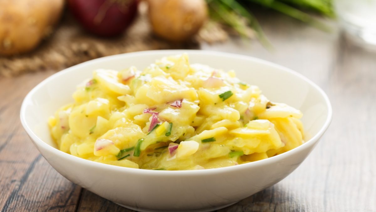 Frontal: ein lauwarmer Kartoffelsalat mit Brühe in einer weißen Schale auf einem rustikalen Untergrund aus Holz. Im Hintergrund sind frische Zwiebeln und Lauch.