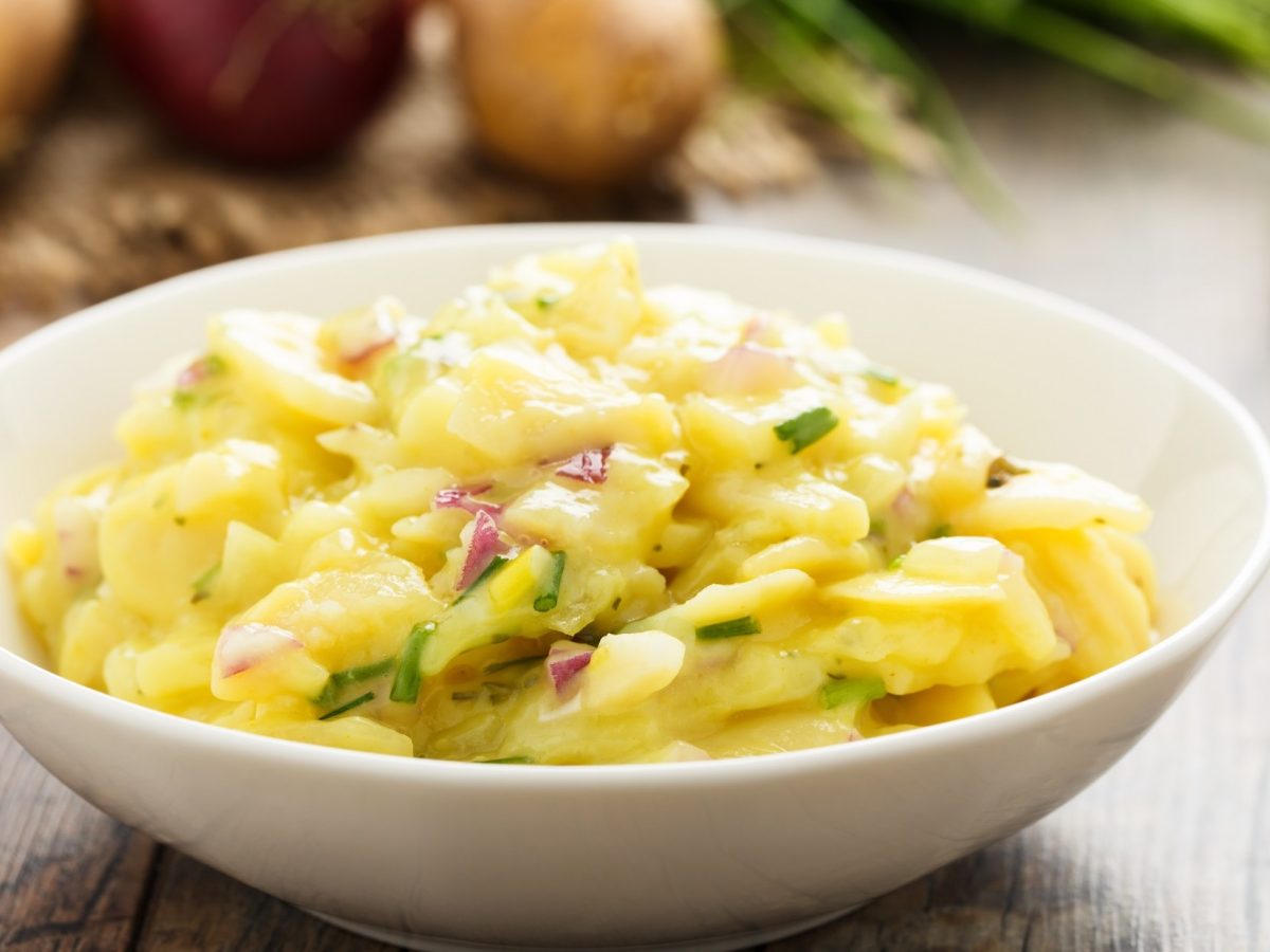 Frontal: ein lauwarmer Kartoffelsalat mit Brühe in einer weißen Schale auf einem rustikalen Untergrund aus Holz. Im Hintergrund sind frische Zwiebeln und Lauch.