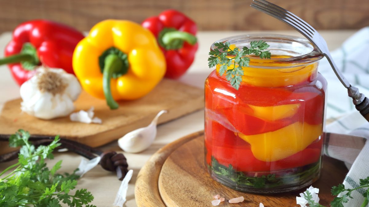 Frontal Ein Glas mit eingelegten Paprika. Im Hintergrund liegen frische rote und gelbe Paprika, Kräuter und Knoblauch. / Paprika einlegen