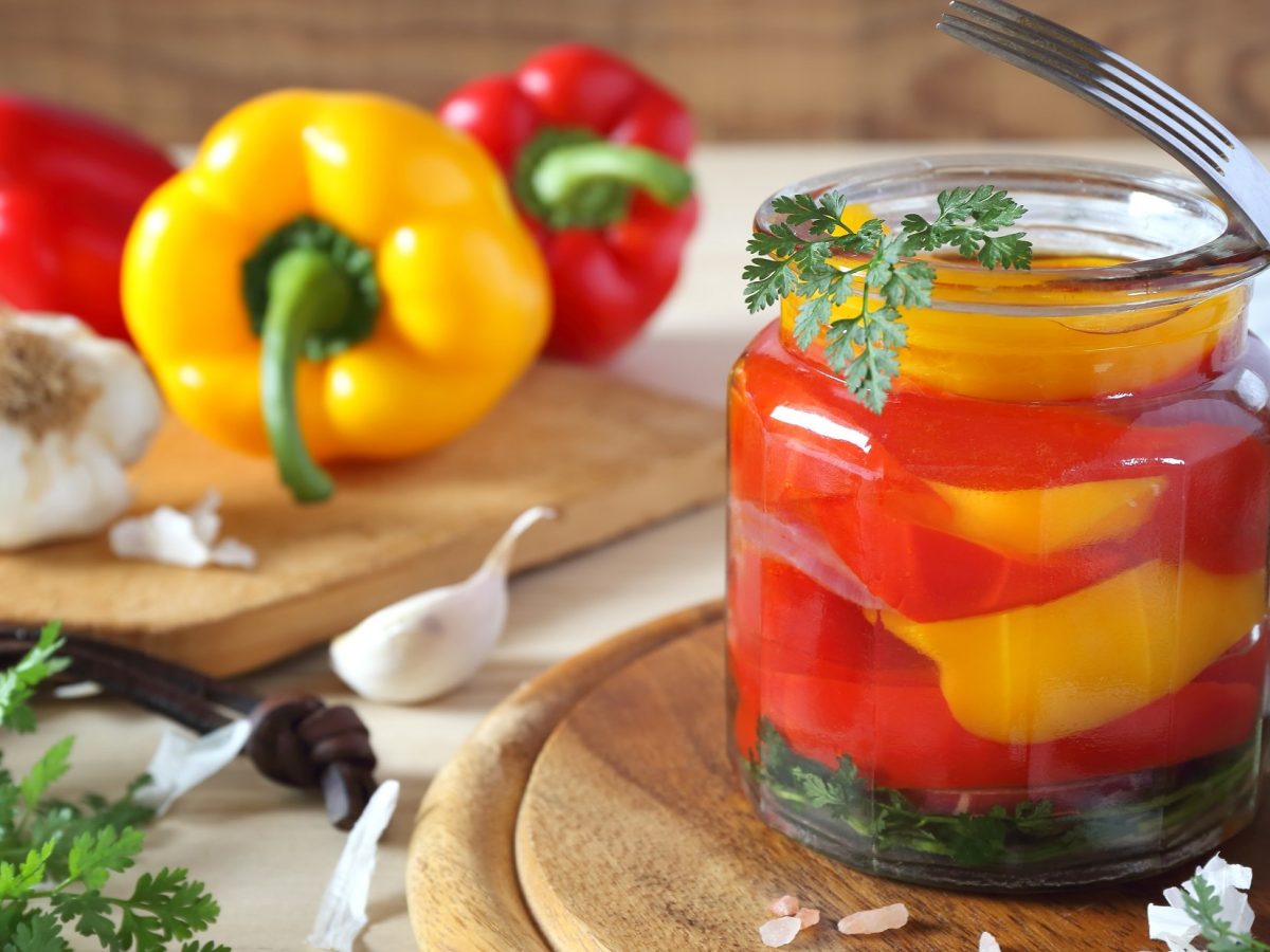 Frontal Ein Glas mit eingelegten Paprika. Im Hintergrund liegen frische rote und gelbe Paprika, Kräuter und Knoblauch. / Paprika einlegen