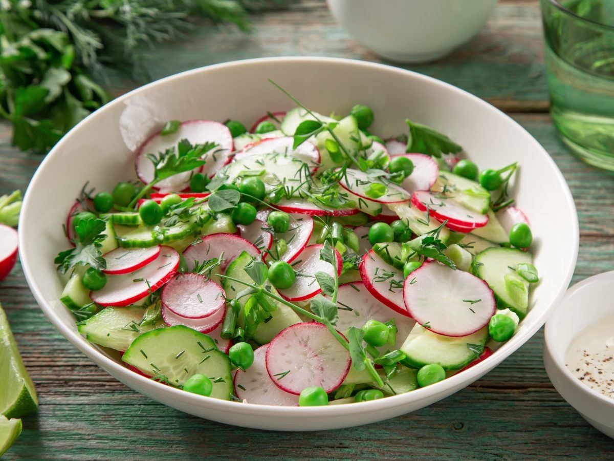 Draufsicht, schräg: Eine weiße Schale Radieschensalat, der nach Omas Rezept zubereitet wurde. Drum herum liege frische Salatzutaten.