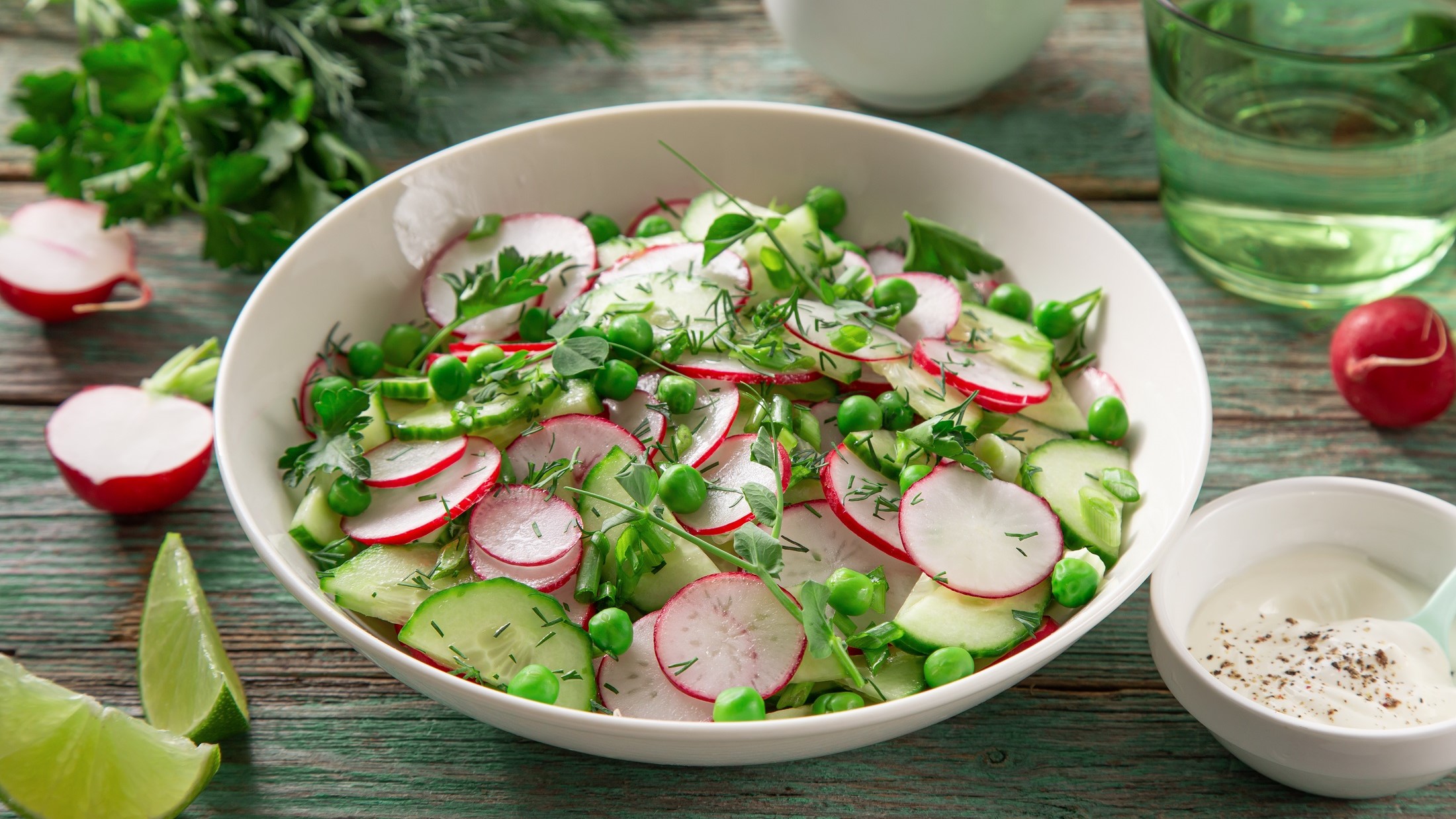 Draufsicht, schräg: Eine weiße Schale Radieschensalat, der nach Omas Rezept zubereitet wurde. Drum herum liege frische Salatzutaten.