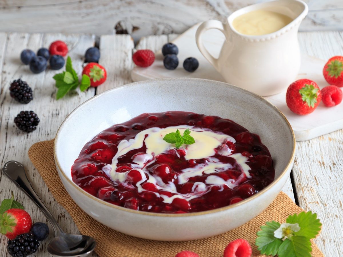 Draufsicht schräg: Eine weiße Schale Rote Grütze mit Vanillesauve auf einem hellen Tisch. Drum herum liegen frische Früchte.