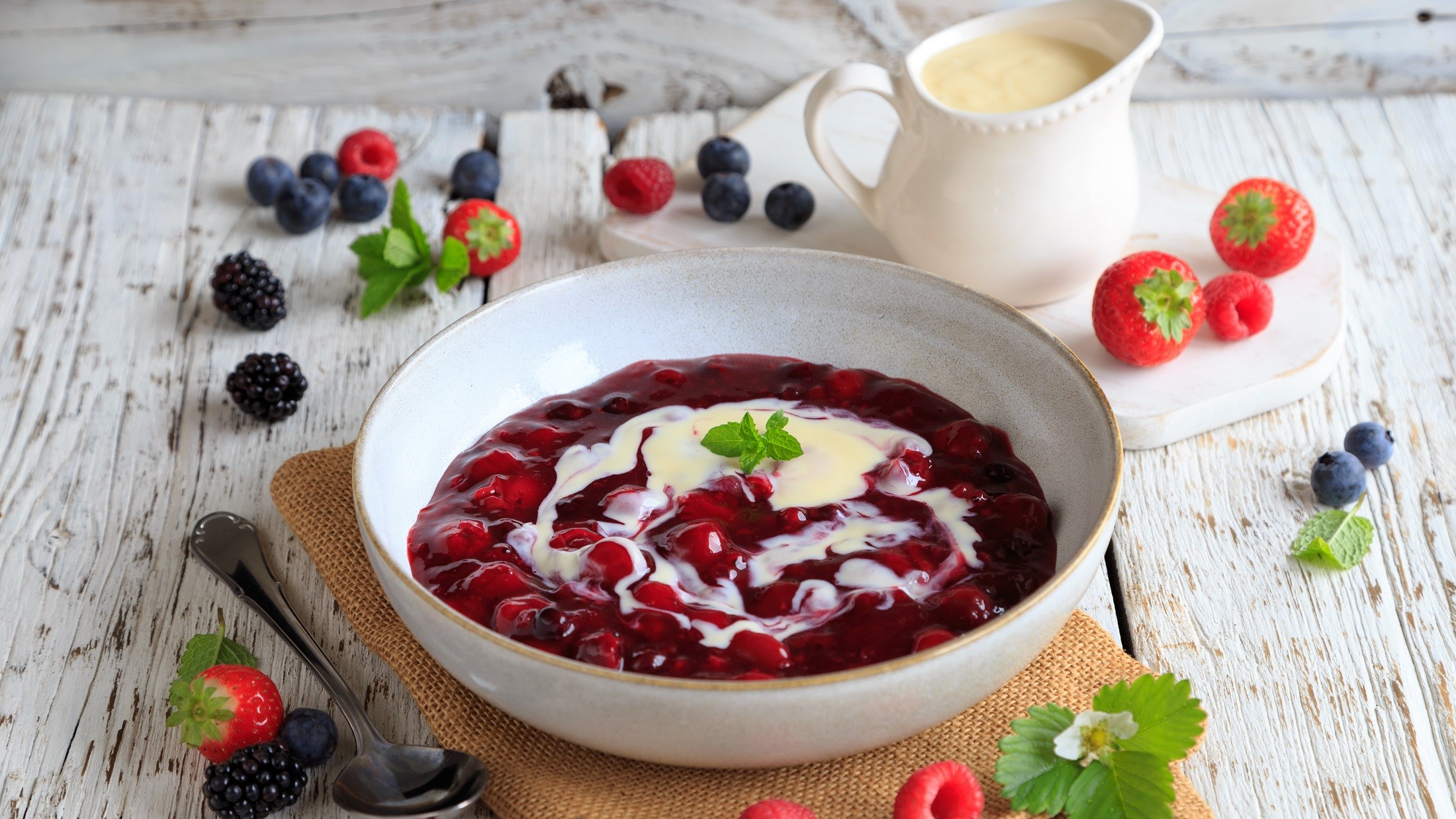 Draufsicht schräg: Eine weiße Schale Rote Grütze mit Vanillesauve auf einem hellen Tisch. Drum herum liegen frische Früchte.