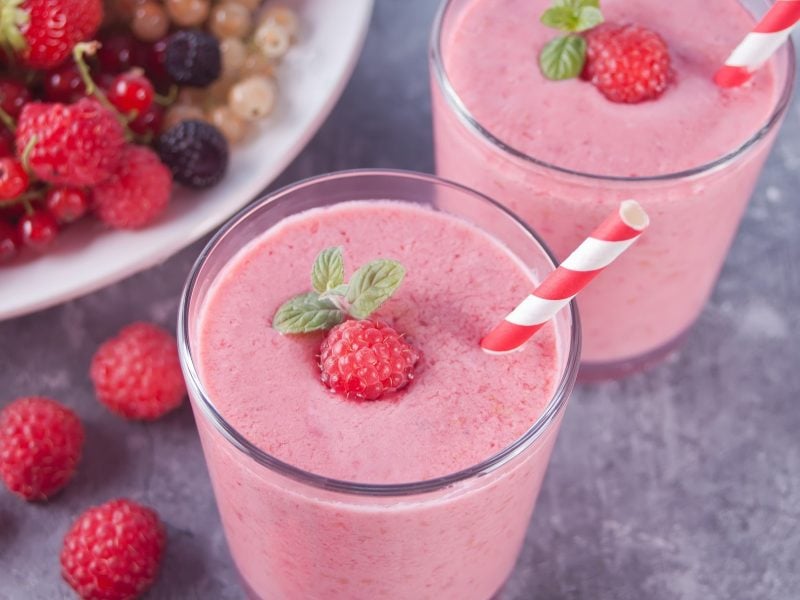 Draufsicht, schräg: Zwei Gläser mit einem Smoothie zum Abnehmen und zwei rot-weiß-gestreiften Strohhalmen. Drum herum und in einer Schüssel liegen frische Beeren, wie etwa Erdbeeren und Johannisbeeren.