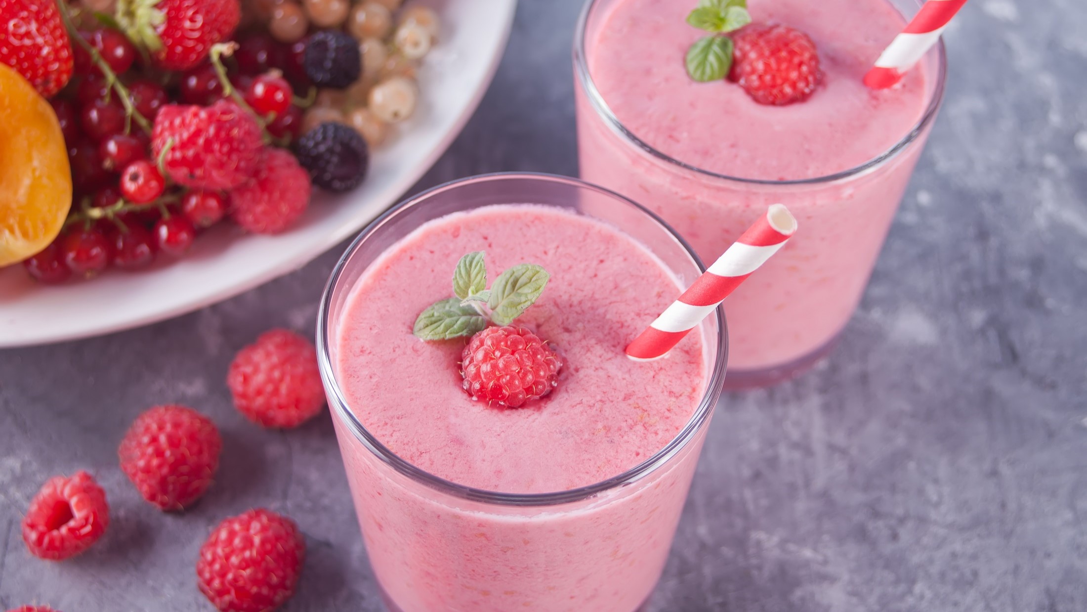 Draufsicht, schräg: Zwei Gläser mit einem Smoothie zum Abnehmen und zwei rot-weiß-gestreiften Strohhalmen. Drum herum und in einer Schüssel liegen frische Beeren, wie etwa Erdbeeren und Johannisbeeren.