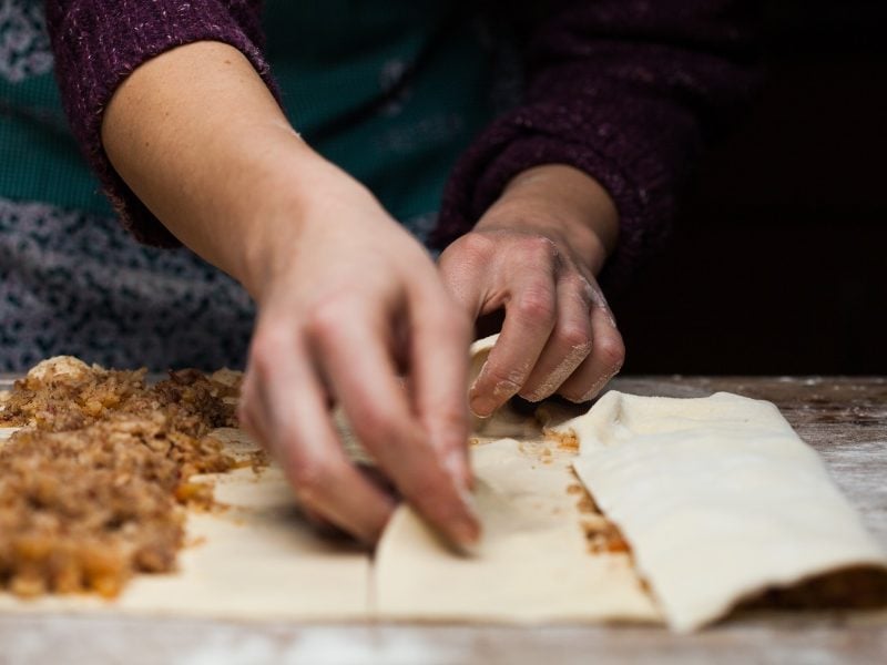 Frontal: Ein Holztisch mit einer dünnen Mehlschicht, auf dem ein ausgezogener Strudelteig liegt. Zwei weibliche Hände die den Strudelteig selbst machen und füllen.