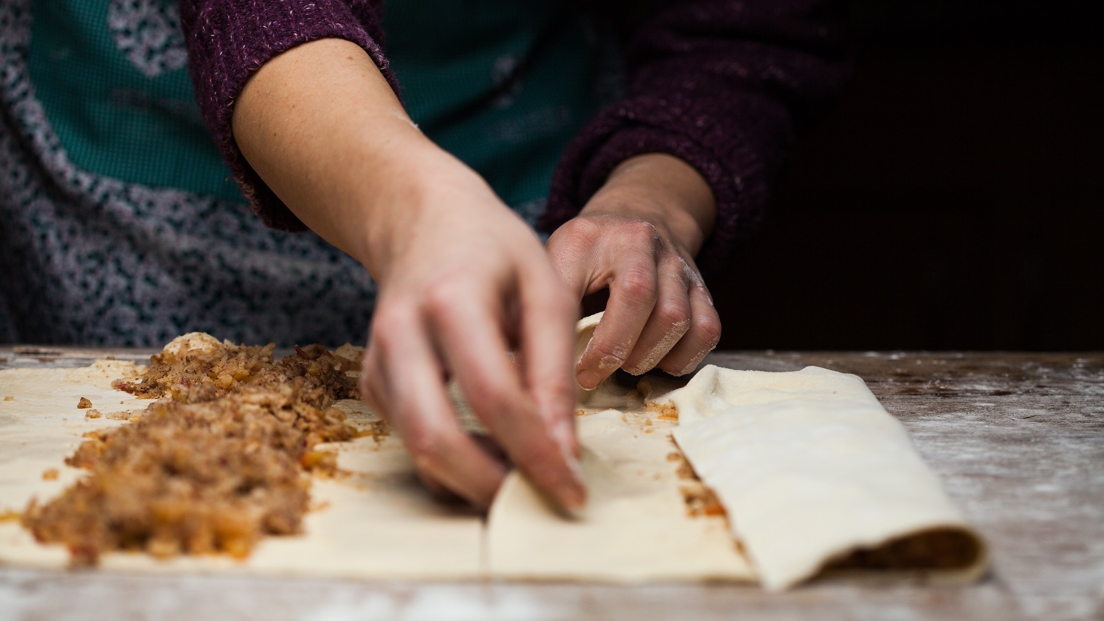 Frontal: Ein Holztisch mit einer dünnen Mehlschicht, auf dem ein ausgezogener Strudelteig liegt. Zwei weibliche Hände die den Strudelteig selbst machen und füllen.