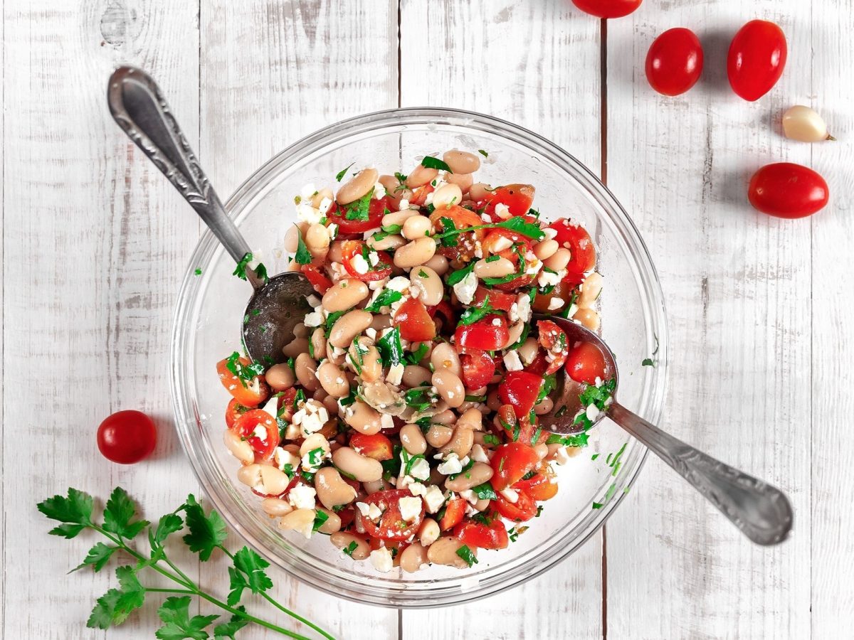 Draufsicht: Ein Teller Weiße-Bohnen-Salat mit Feta auf einem weißem Untergrund aus Holz. Drumherum liegen frische Tomaten und frische Petersilie.