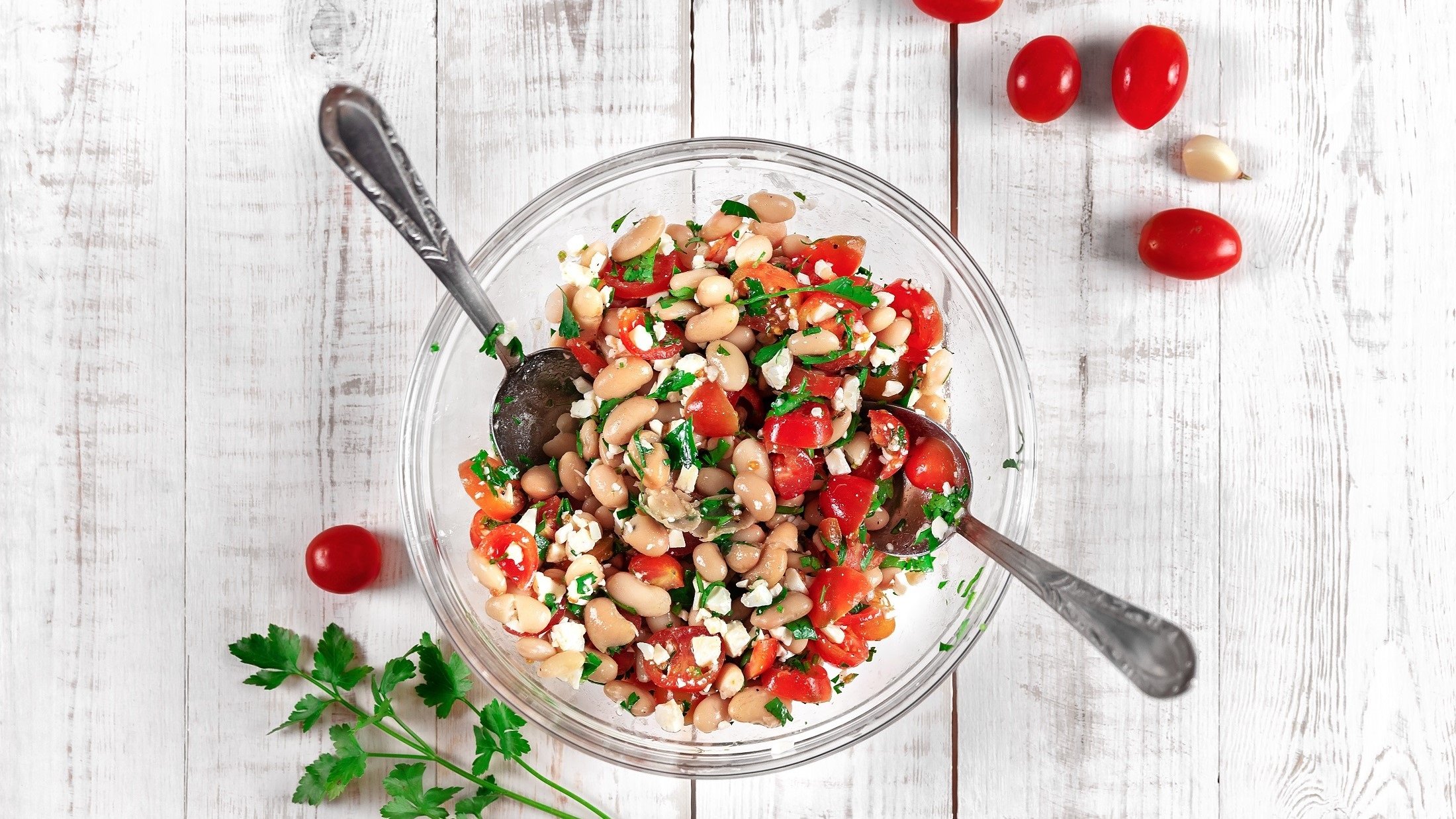 Draufsicht: Ein Teller Weiße-Bohnen-Salat mit Feta auf einem weißem Untergrund aus Holz. Drumherum liegen frische Tomaten und frische Petersilie.