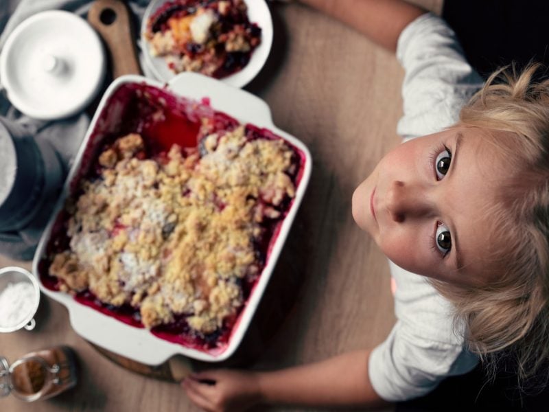 Draufsicht. Dunkle Anmutung: Ein Kind schaut nach oben in die Kamera. Es steht vor einem Tisch auf dem Backzutaten sowie eine Auflaufform mit einem Zwetschgen-Crumble liegen.