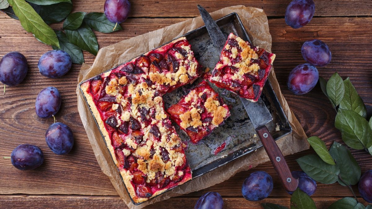 Draufsicht: Ein Zwetschgenkuchen vom Blech auf einem rustikalen Untergrund aus Holz. Drum herum liegen frische Zwetschgen und grüne Blätter.