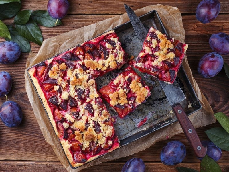 Draufsicht: Ein Zwetschgenkuchen vom Blech auf einem rustikalen Untergrund aus Holz. Drum herum liegen frische Zwetschgen und grüne Blätter.