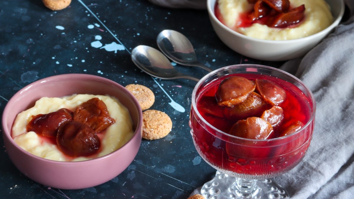 Draufsicht: Zwei Schalen Pudding mit Zwetschgenröster als Sauce. Daneben eine Schale aus Glas mit Zwetschgenröster-Kompott. Alles steht auf einem rustikalen Tisch. Daneben liegen bei Löffel und zwei Kekse,