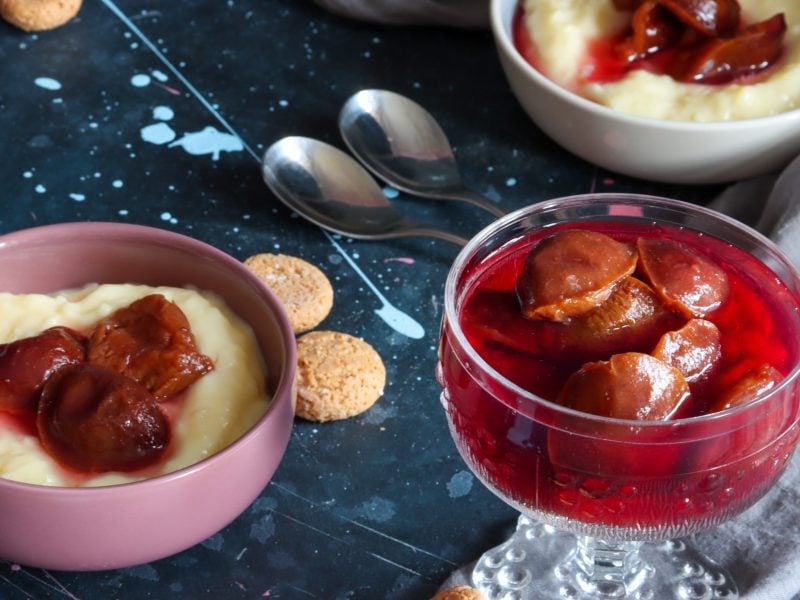 Draufsicht: Zwei Schalen Pudding mit Zwetschgenröster als Sauce. Daneben eine Schale aus Glas mit Zwetschgenröster-Kompott. Alles steht auf einem rustikalen Tisch. Daneben liegen bei Löffel und zwei Kekse,