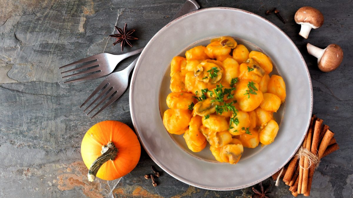 Gnocchi mit Kürbissauce und Champignons auf einem weißen Teller, daneben ein Kürbis und Gewürze, von oben fotografiert.