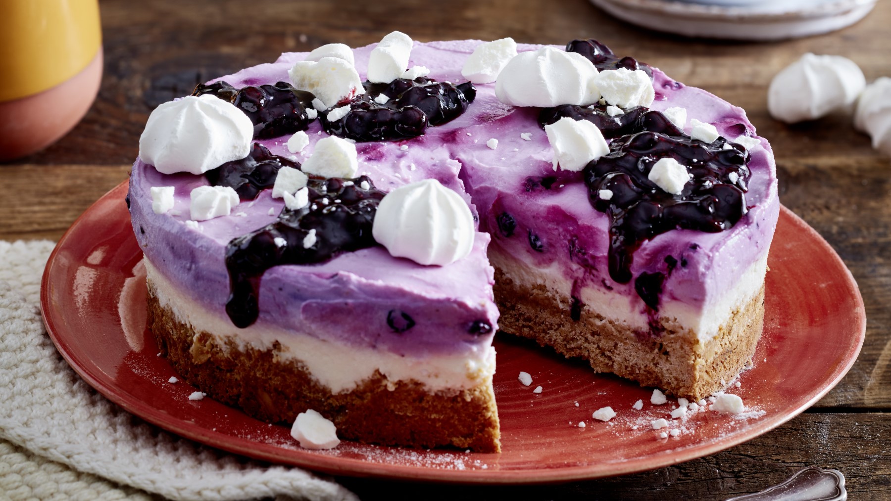 Angeschnittene Heidelbeer-Skyr-Torte mit Baiser auf rotem Teller.