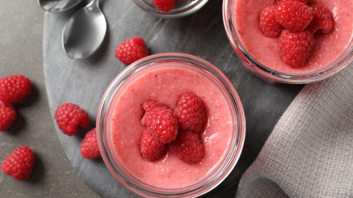 Draufsicht: Zwei Gläser mit veganer Himbeer-Mousse und frischen Himbeeren darauf und daneben, grauer Untergrund.