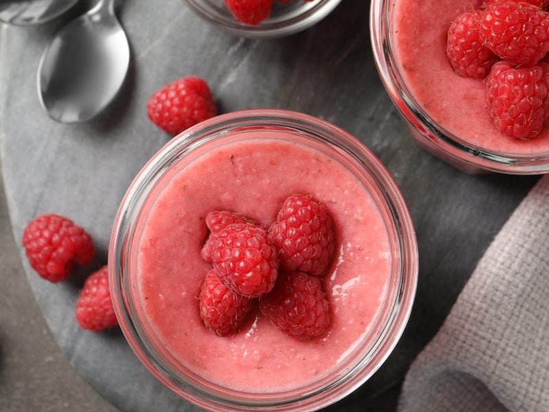 Draufsicht: Zwei Gläser mit veganer Himbeer-Mousse und frischen Himbeeren darauf und daneben, grauer Untergrund.