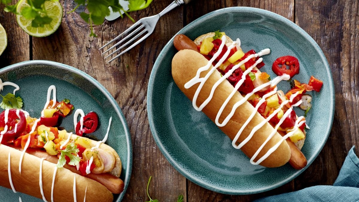 Draufsicht: Zwei Teller Hotdog mit Ananas & Jalapeños und Sauce drüber.