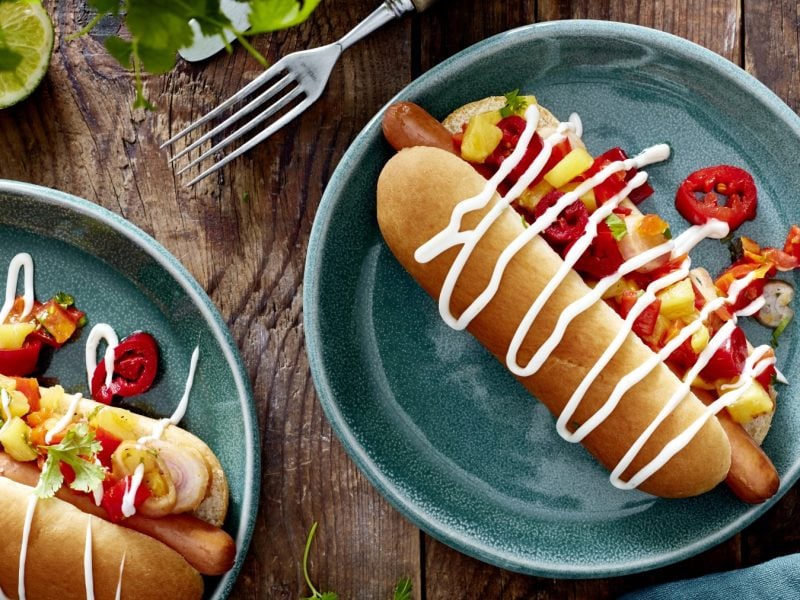 Draufsicht: Zwei Teller Hotdog mit Ananas & Jalapeños und Sauce drüber.