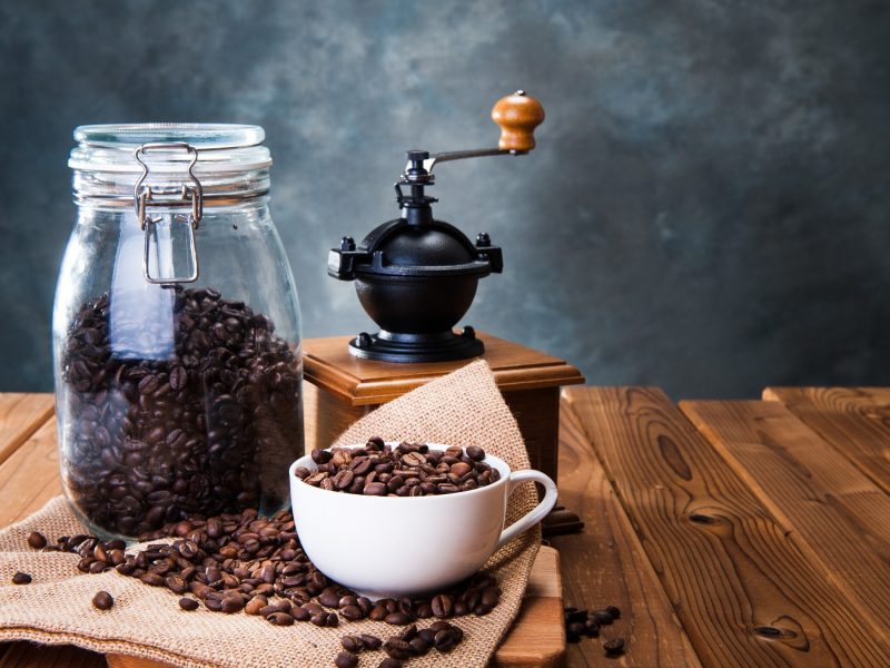 Frische Kaffeebohnen in Glasbehälter und Tasse, arrangiert neben einer schwarzen Hand-Kaffeemühle auf Holz.