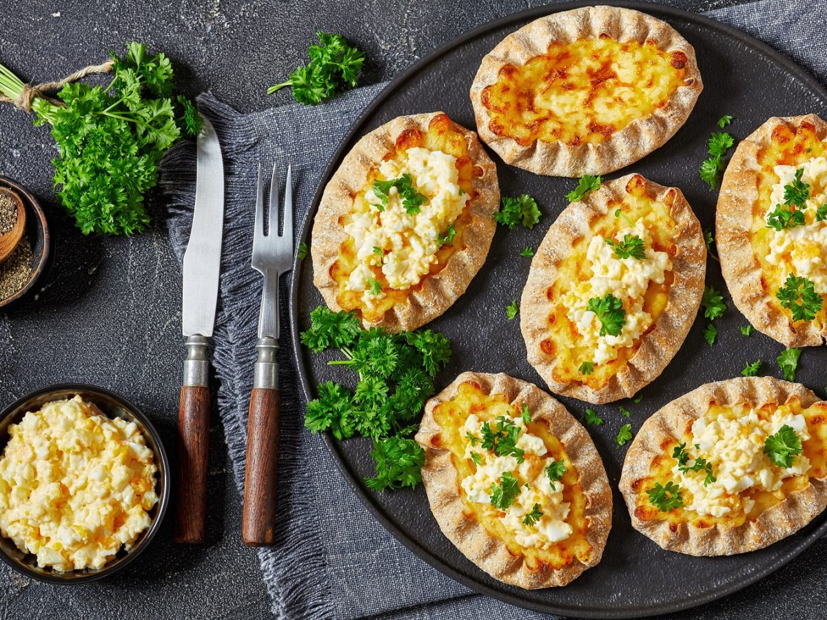 Karelische Piroggen, mit Ei-Butter bestrichen von oben fotografiert, daneben Schale mit Ei-Butter, Messer und Gabel sowie frische Kräuter und Gewürze.