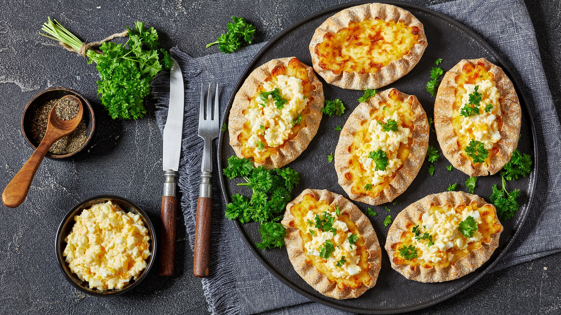 Karelische Piroggen, mit Ei-Butter bestrichen von oben fotografiert, daneben Schale mit Ei-Butter, Messer und Gabel sowie frische Kräuter und Gewürze.