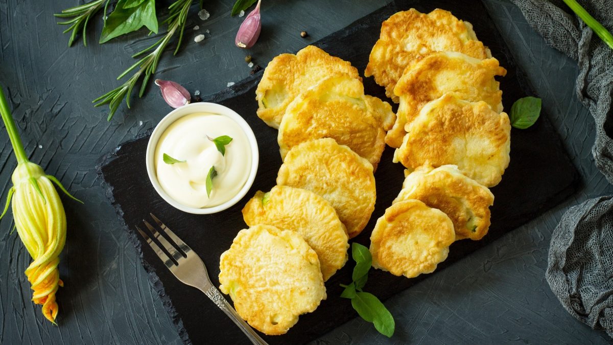 Draufsicht auf Kolokithakia tiganita auf einer schwarzen Steinplatte, daneben ein Dip und eine Zucchiniblüte.