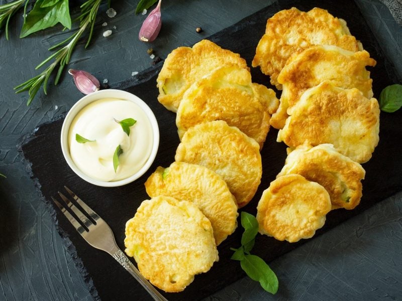 Draufsicht auf Kolokithakia tiganita auf einer schwarzen Steinplatte, daneben ein Dip und eine Zucchiniblüte.