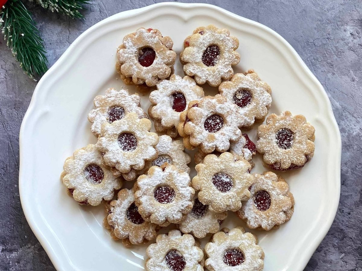 Die Linzer Augen auf einem weißen Teller und grauem Steintisch mit etwas Weihnachtsdekoration direkt von oben fotografiert.