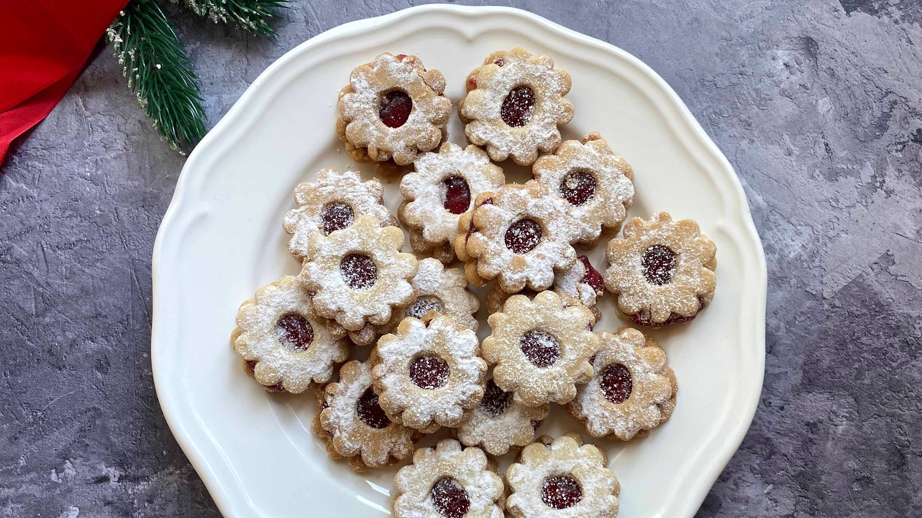 Die Linzer Augen auf einem weißen Teller und grauem Steintisch mit etwas Weihnachtsdekoration direkt von oben fotografiert.