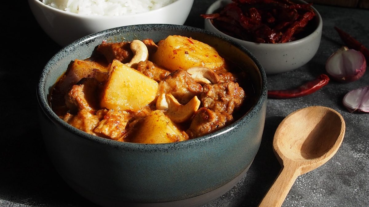 Eine schwarze Schale mit dem Massaman Curry auf einem grauen Steintisch von schräg oben fotografiert. Im hIntegrund unscharfv Reis und eine schale mit getrockneten Chilis zu erkennen.