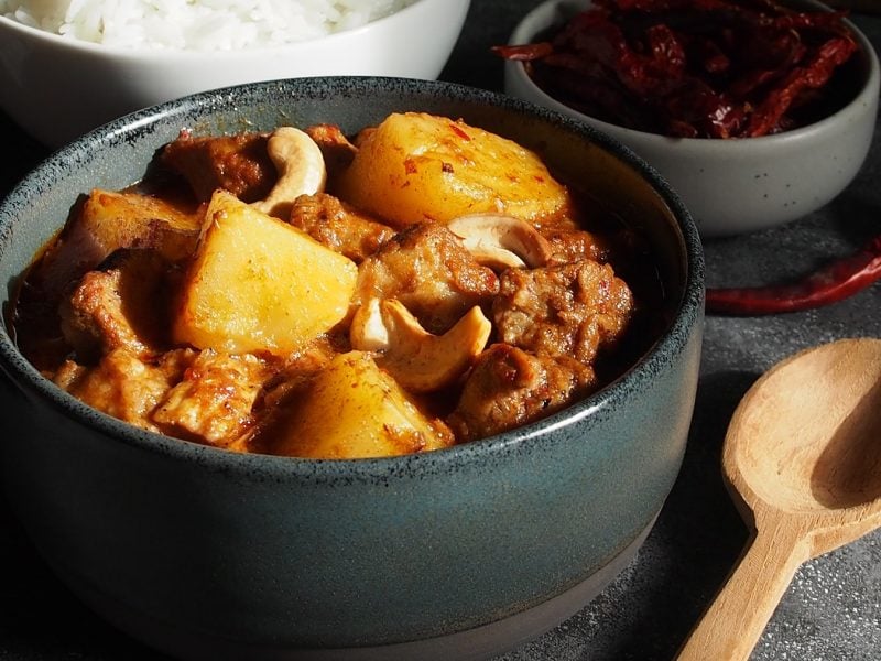 Eine schwarze Schale mit dem Massaman Curry auf einem grauen Steintisch von schräg oben fotografiert. Im hIntegrund unscharfv Reis und eine schale mit getrockneten Chilis zu erkennen.