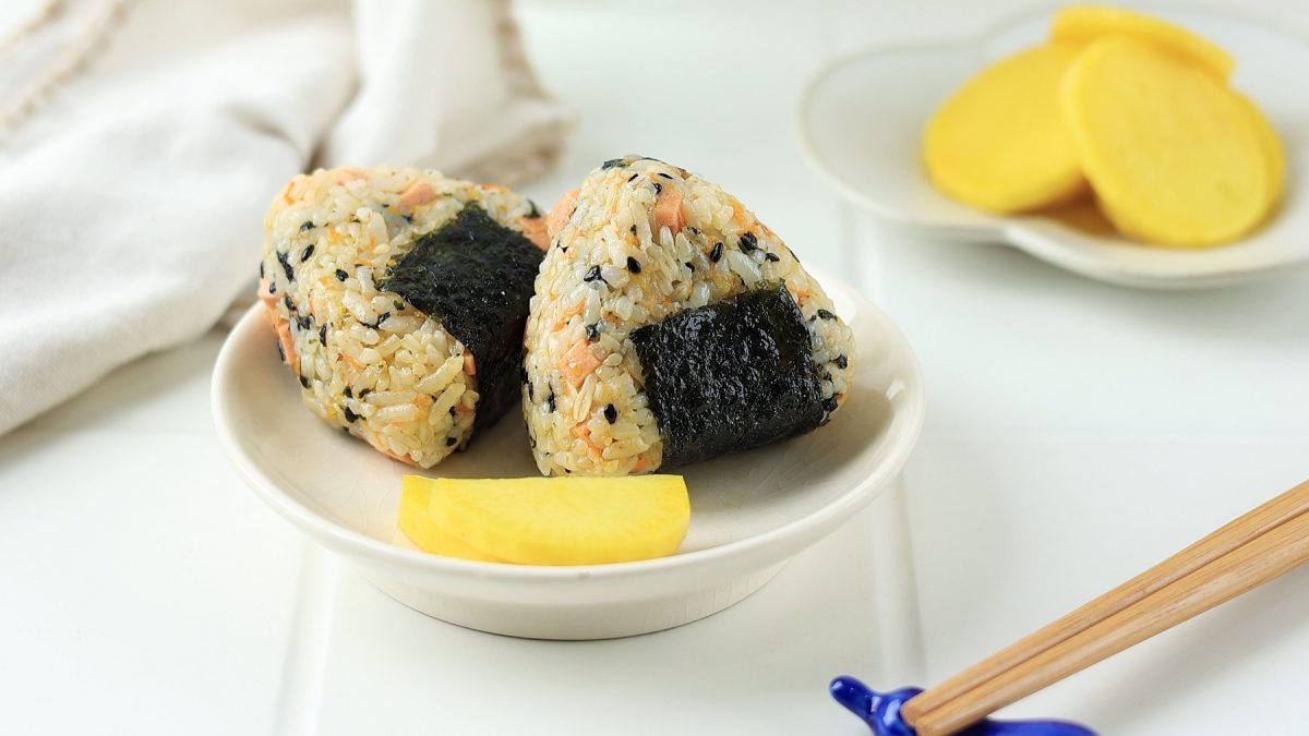 Ein kleiner weißer Teller mit zwei Onigiri un dgelbem Rettich in Scheiben geschnitten auf weißem Untergrund und beiger Tischdecke. Alles von der Seite und leicht schräg oben fotografiert.