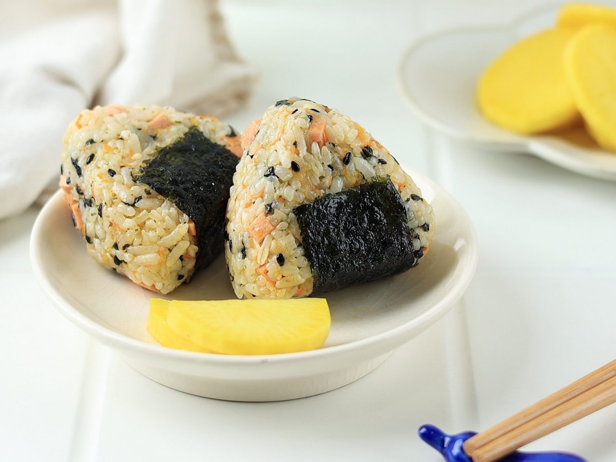 Ein kleiner weißer Teller mit zwei Onigiri un dgelbem Rettich in Scheiben geschnitten auf weißem Untergrund und beiger Tischdecke. Alles von der Seite und leicht schräg oben fotografiert.