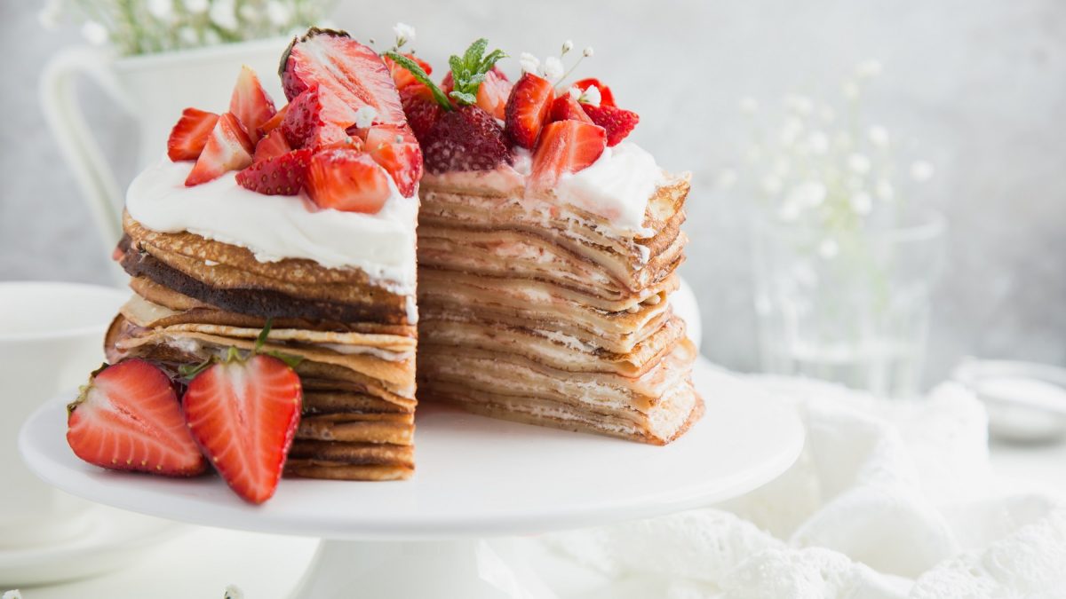 Die Pfannkuchen-Torte auf einer weißen Tortenplatte vor hellem Hintergrund und auf einem weißen Tisch. Alles von der Seite fotografiert.